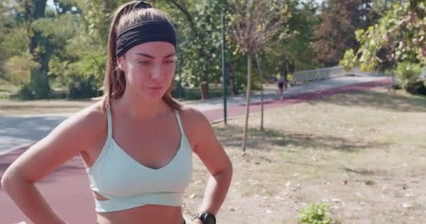 Femme déterminée en tenue de sport prenant une pause de jogging. Une coureuse reprenant son souffle avec une toile de fond de parc sereine. - Séquence, vidéo