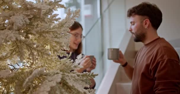 A young couple shares a cheerful moment with hot beverages by a snowy Christmas tree, embodying the festive holiday spirit and creating warm memories together. - Footage, Video