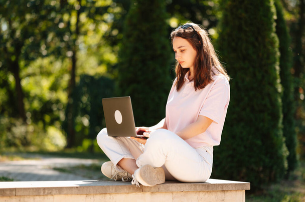 Nuori keskittynyt nainen istuu puistossa käyttäen kannettavaa tietokonetta tutkimuksiinsa aurinkoisena päivänä.. - Valokuva, kuva