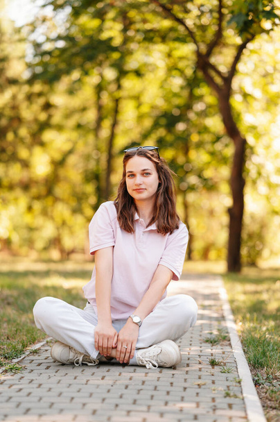 Młoda wymarzona studentka siedzi w parku na chodniku w słoneczny dzień lata. - Zdjęcie, obraz