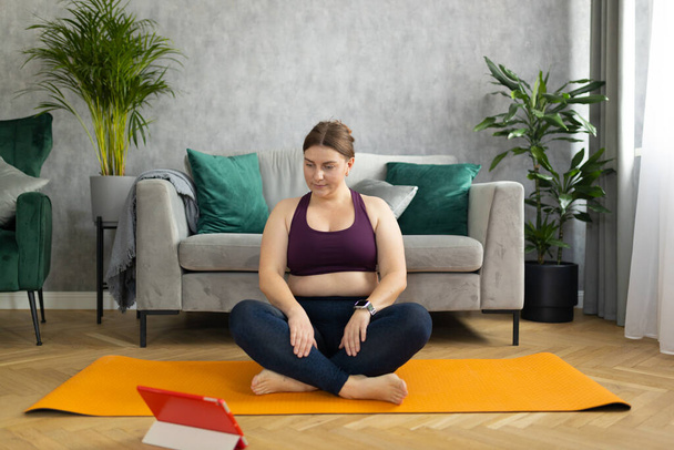 Sporty plus size woman in sportswear working out at home. Sports, activity and weight loss concept. Young plus size woman in leggings exercising on the mat, watching online video via tablet. - Foto, Imagen