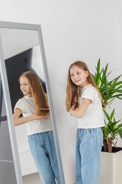 Mosolygó kiskorú gyermek fésülködik hosszú haját, miközben nézi a tükörképét teljes hosszúságú tükörben fényes szobában. - Fotó, kép