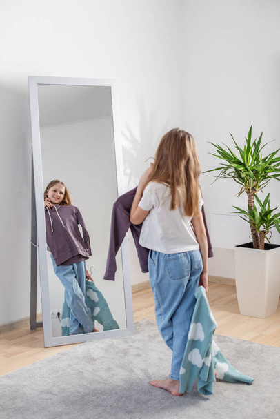 Fille examine ses options de tenue reflétée dans un miroir pleine longueur, entouré d'une ambiance de chambre confortable avec plante d'intérieur. - Photo, image