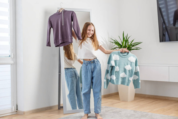 Een glimlachend meisje tilt een hanger met een trui hoog als ze kiest haar outfit in de voorkant van een slaapkamer spiegel. - Foto, afbeelding