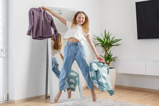 Χαρούμενη κοπέλα χορεύει χαρούμενα στο δωμάτιό της, κρατώντας ψηλά δύο πουλόβερ, με την παιχνιδιάρικη αντανάκλασή της σε ολόσωμο καθρέφτη. - Φωτογραφία, εικόνα