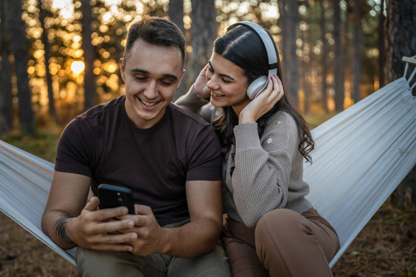 Homme et femme jeune couple adulte dans la nature écouter de la musique sur casque utiliser un téléphone portable smartphone ou faire une application d'appel vidéo - Photo, image