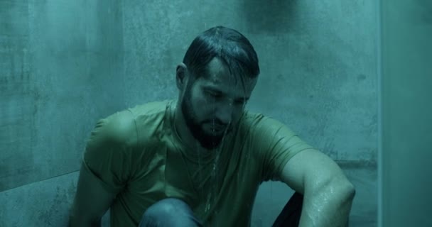 Zdenerwowany brodaty dorosły mężczyzna w ubraniu casual siedzi pod prysznicem w łazience w domu - Materiał filmowy, wideo
