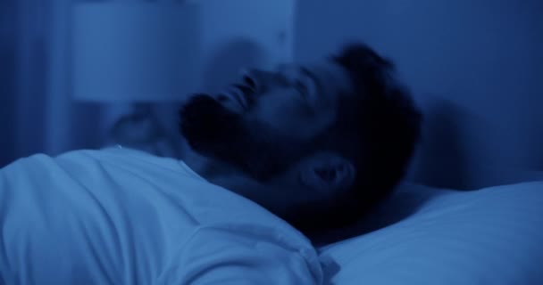 Reaaliaikainen näkymä parrakas aikuinen mies kärsii unettomuus etsii ylös, kun makaa selkä kääntöpuolella sängyllä ja pysty nukkumaan yöllä huoneessa - Materiaali, video