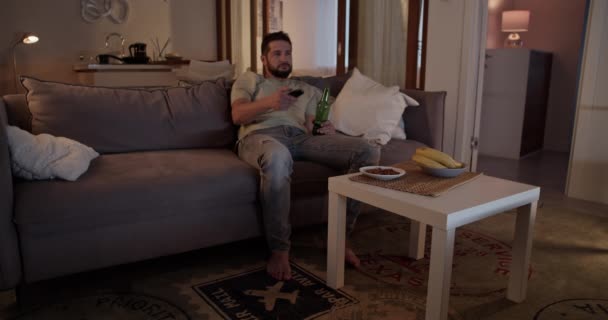 Homme adulte barbu déprimé en vêtements décontractés tenant télécommande regarder la télévision tout en buvant de la bière assis sur le canapé dans le salon dans l'appartement - Séquence, vidéo