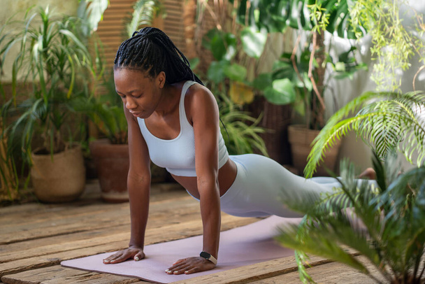 Aktive Afroamerikanerin dehnt sich mit geschlossenen Augen auf einer Fitnessmatte, die von tropischen Zimmerpflanzen umgeben ist. Sportliche schwarze Frauen machen Yoga, entwickeln Beweglichkeit für einen gesunden Körper im gemütlichen Sportstudio. - Foto, Bild