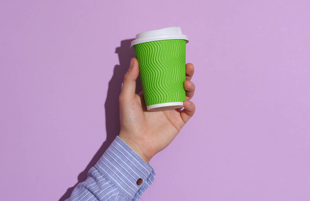 Mužská ruka v košili drží zelený karton šálek kávy na fialové pastelové pozadí se stínem - Fotografie, Obrázek