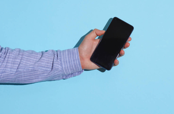Männliche Hand im Hemd hält Smartphone auf blauem Hintergrund mit Schatten - Foto, Bild