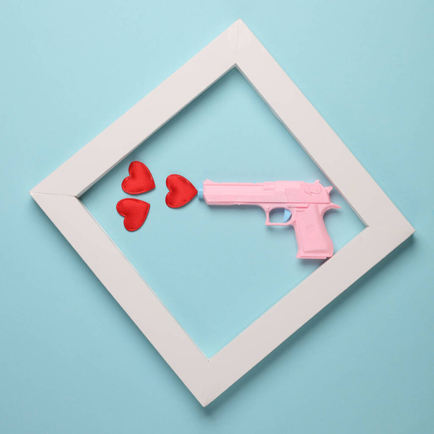 Kunststoffpistole mit Herzen in weißem Rahmen auf blauem Hintergrund. Minimalismus. Kreatives Layout. Liebeskonzept - Foto, Bild
