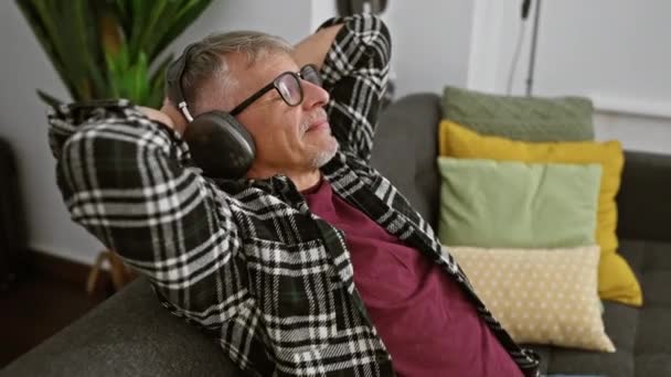 Rento vanhempi mies nauttii musiikkia kuulokkeet, makuuasennossa kotona sohvalla ympäröivät värikkäät tyynyt. - Materiaali, video