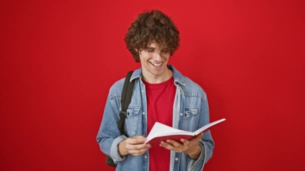 Glimlachende jongeman met krullend haar die een boek leest tegen een levendige rode achtergrond. - Video