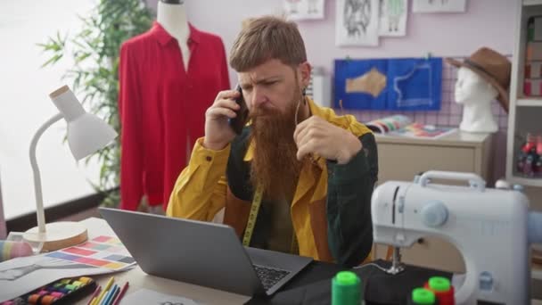 Rudowłosy mężczyzna z brodą pracuje w sklepie krawieckim rozmawiając przez telefon i używając laptopa. - Materiał filmowy, wideo