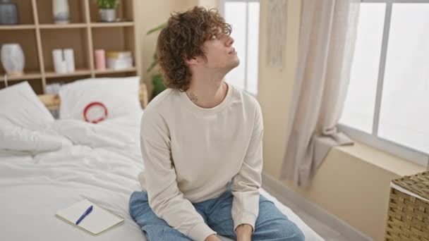 Een peperdure jongeman met krullend haar zit op een bed in een lichte, gezellige slaapkamer en straalt een sfeer van bezinning uit. - Video
