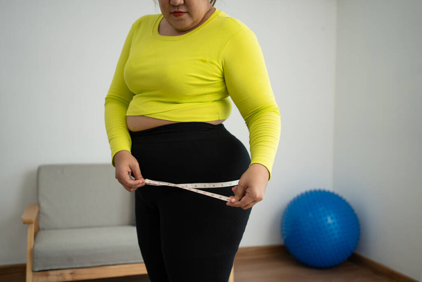 Ylipainoinen nainen, joka käyttää mittanauhaa vyötärön ja vatsan rasvan ympärillä, lihava aasialainen nainen treenaa kotona. Käsite terveellistä, ruokaa terveydelle ja elämäntavalle, laihtuminen - Valokuva, kuva