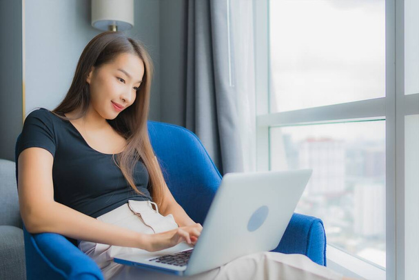 Portré gyönyörű fiatal ázsiai nő használja laptop kanapén a nappali területén - Fotó, kép