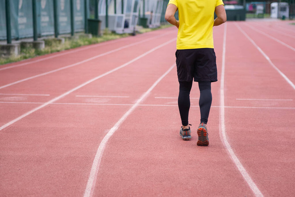 z tyłu widok człowieka w żółtej koszulce bieganie i jogging wzdłuż toru tętniącego życiem stadionu, współczesnej hali sportowej w parku miejskim. - Zdjęcie, obraz
