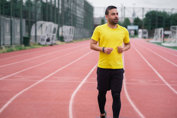 Energetischer junger Mann joggt auf einer leeren Strecke mit rotem Gummibelag und knackig weißen Markierungen auf einem Sportplatz im Stadtpark, der für eine gesunde Lebensweise steht.  - Foto, Bild