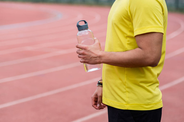 athlete man takes a moment to catch his breath on the running track, holding refreshing water bottle at the city sports arena.  - Photo, Image