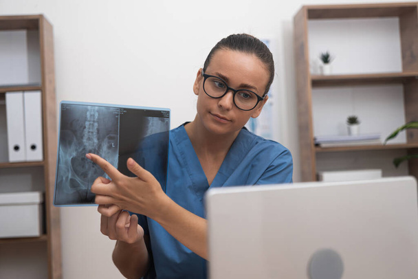Arzt in blauer Uniform zeigt mit dem Finger auf Röntgenfilm, der dem Patienten im Laptop während der Online-Telemedizin-Beratung gezeigt wird - Foto, Bild