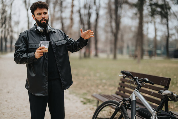 Професійний чоловік-підприємець відпочиває в парку зі своїм велосипедом, виступаючи на бездротовій гарнітурі, тримаючи чашку кави. - Фото, зображення