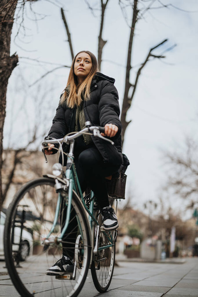 Młoda dorosła kobieta w zimowym płaszczu pewnie jeździ rowerem po miejskiej ścieżce, emanując poczuciem wolności i aktywnym stylem życia.. - Zdjęcie, obraz