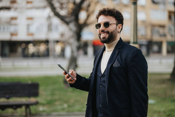 Hyvin pukeutunut mies seisoo matkapuhelimeensa uppoutuneessa kaupunginpuistossa, jossa vallitsee teknologian ja luonnon tasapaino.. - Valokuva, kuva