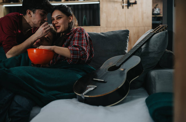 Ομάδα νεαρών φίλων έχουν μια ζεστή συγκέντρωση στο σπίτι με μια κιθάρα και μπολ με σνακ στον καναπέ - Φωτογραφία, εικόνα
