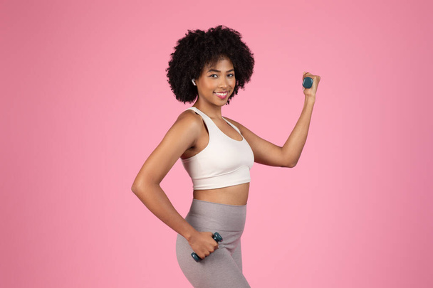Uśmiechnięta Afroamerykanka w odzieży sportowej pokazuje swój biceps, pokazuje siłę i szczęście podczas treningu - Zdjęcie, obraz
