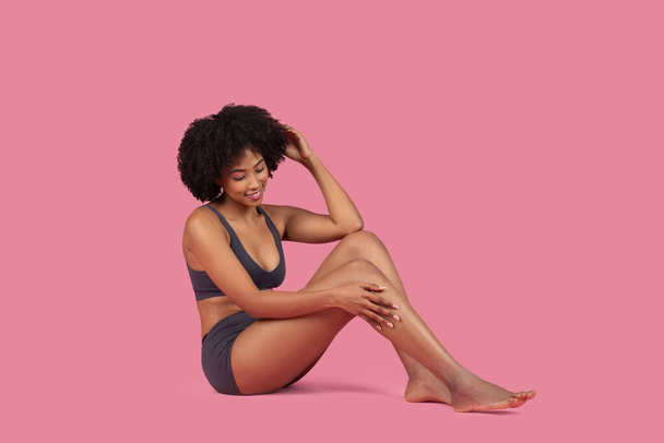 Une jolie jeune femme afro-américaine aux cheveux bouclés courts portant des sous-vêtements assis sur un fond rose, touchant ses cheveux, regardant vers le bas avec un sourire doux - Photo, image