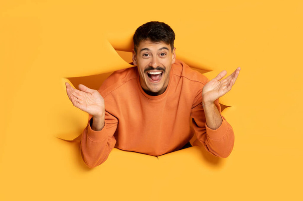Έκπληκτος αστείος άνθρωπος σκάει μέσα από μια τρύπα σε ένα κίτρινο φόντο, με ένα παιχνιδιάρικο και ενθουσιασμένοι έκφραση - Φωτογραφία, εικόνα