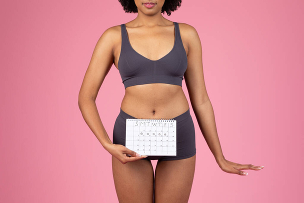 Una giovane donna afroamericana in reggiseno sportivo tiene un calendario del ciclo mestruale contro il suo basso addome su uno sfondo rosa - Foto, immagini