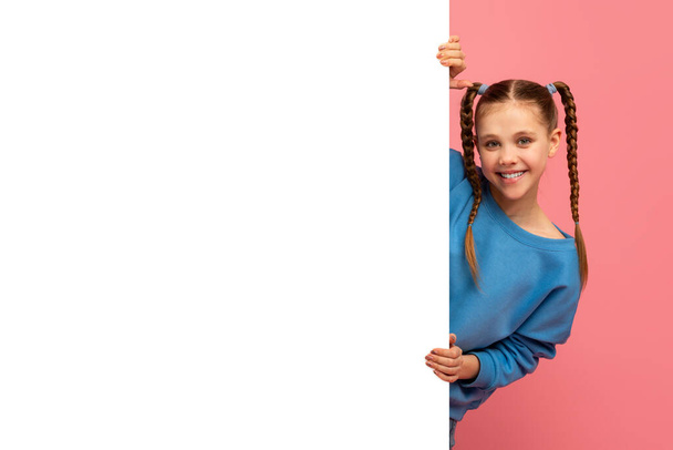 Lächelndes junges Mädchen mit geflochtenem Haar, das hinter einer leeren weißen Tafel vor rosa Hintergrund hervorlugt - Foto, Bild