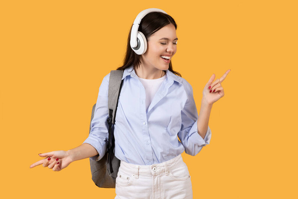 Une jeune femme joyeuse en tenue décontractée, appréciant la musique avec écouteurs sur l'oreille, dansant avec un fond jaune - Photo, image