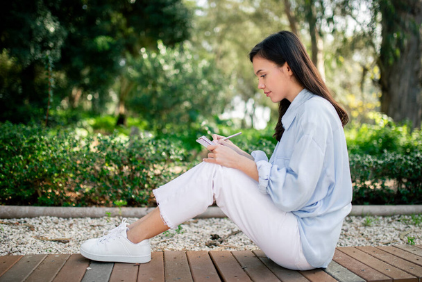 Mladá žena sedí na zemi v klidném prostředí parku, zaujatá svým chytrým telefonem, obklopená bujnou zelení - Fotografie, Obrázek