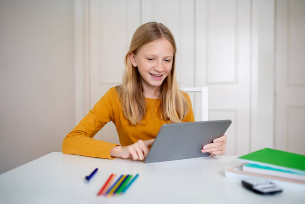 Fröhliches Teenie-Mädchen, das zu Hause mit einem digitalen Tablet beschäftigt ist, glückliche Teenagerin, die modernes Gerät für Studium oder Freizeit nutzt und drinnen am Schreibtisch sitzt - Foto, Bild