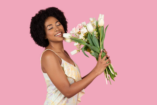 A joyful African American woman with curly hair gently hugs a bouquet of white tulips and other flowers, eyes closed in delight, against a pink background - Photo, Image
