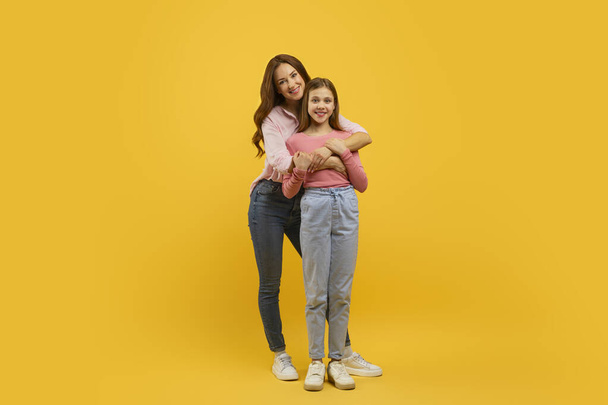 Δύο χαμογελαστές γυναίκες ένας ενήλικας και ένα παιδί, μοιράζονται μια στοργική αγκαλιά σε ένα ζωντανό κίτρινο φόντο - Φωτογραφία, εικόνα