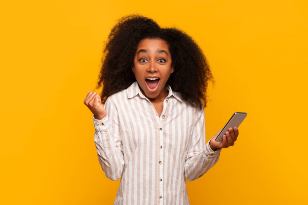 Lächelnde junge schwarze Dame begeistert von dem, was sie auf ihrem Handybildschirm vor gelbem Hintergrund sieht - Foto, Bild