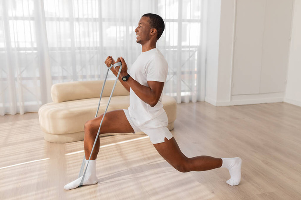Активний афроамериканський чоловік використовує групу опору, щоб відпрацювати ноги в легкому сучасному просторі - Фото, зображення