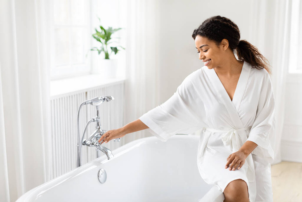 Mujer afroamericana en una lujosa bata blanca está alcanzando el grifo, elementos elegantes destacan el baño moderno - Foto, Imagen
