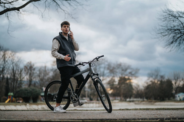 Rento teini pitää taukoa pyöräilystä puhuakseen älypuhelimellaan rauhallisessa puistossa.. - Valokuva, kuva
