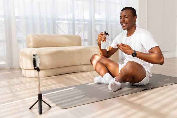 Afroamerikaner lächelt, als er ein Video mit seinem Handy aufzeichnet, das auf einem Stativ montiert ist und in einem gut beleuchteten Wohnzimmer auf einer Yogamatte sitzt - Foto, Bild