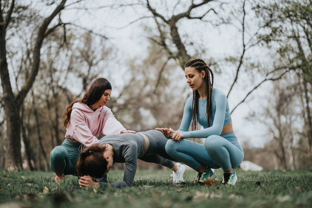 Una sesión de entrenamiento de fitness se lleva a cabo en un parque, con dos mujeres ayudando a un compañero en la realización de tablones. - Foto, Imagen
