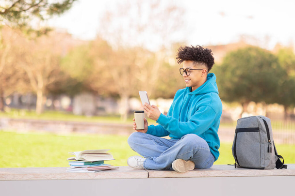 リラックスしたブラジルの男の学生は,キャンパスでコーヒーブレイクを楽しんでいる間,スマートフォンをチェックし,近くの本で座っています - 写真・画像