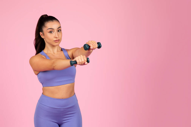 A strong young woman athlete in blue workout attire focused on lifting dumbbells against a pink background - Photo, Image