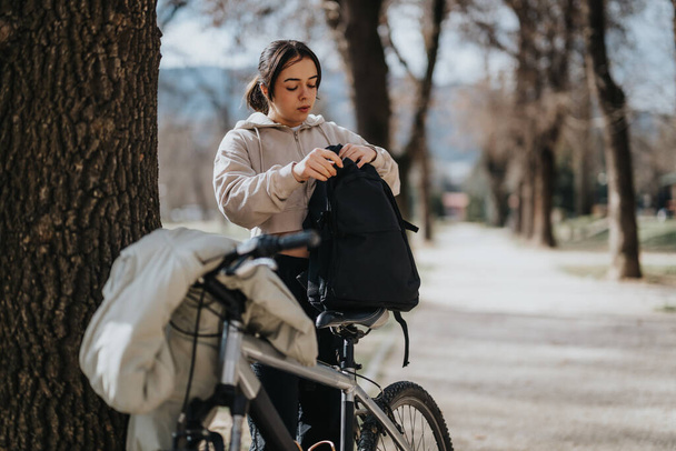 Gondtalan fiatal nő szünetet tart a biciklijével a napos parkban, turkál a hátizsákjában.. - Fotó, kép